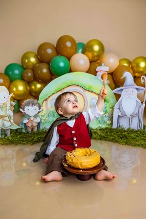 Toddler dressed as Hobbit sits with yellow smash cake, surrounded by Lord of the Rings themed balloons and character cutouts.