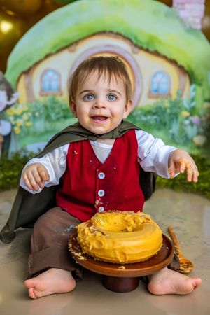 Smiling baby in hobbit costume celebrating first birthday with yellow smash cake in front of Bag End backdrop.