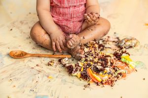 Baby in red romper sitting on floor smashing birthday cake, covered in chocolate crumbs and frosting.