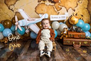 Baby boy Rowan celebrates his first birthday with a travel-themed cake smash, surrounded by balloons, a toy plane, and vintage suitcases.