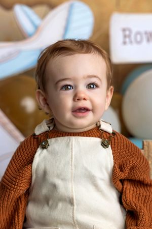 Adorable baby boy named Row wearing brown knit sweater and cream overalls, smiling at camera during first birthday photo.
