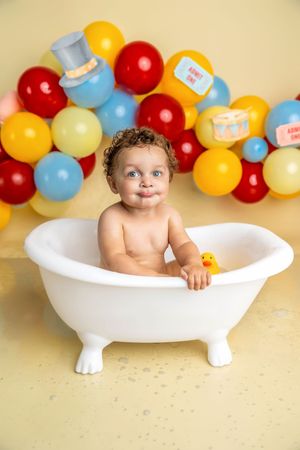 Smiling baby sitting in a white clawfoot bathtub with a rubber duck, surrounded by colorful balloons.