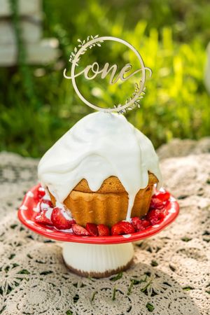 A white-frosted bundt cake topped with a "one" cake topper, decorated with strawberries on a red cake stand outdoors.