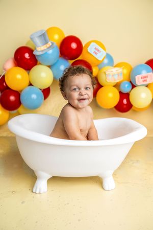 Smiling baby sitting in a white mini bathtub surrounded by colorful red, yellow, and blue balloons on a yellow background.