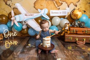 Baby boy celebrates his first birthday with a cake smash, surrounded by travel-themed balloons, a toy airplane, and vintage suitcases.