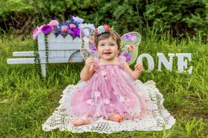 Baby girl in pink fairy dress with wings celebrates first birthday outdoors with floral backdrop and ONE sign.