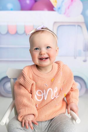 Smiling baby girl in pink 'one' sweater and bow headband sitting in white chair for first birthday photo.
