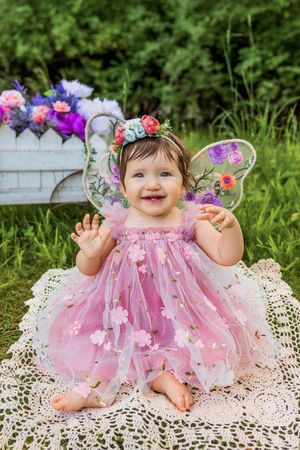 Smiling baby girl in pink fairy dress with butterfly wings and floral crown sits on lace blanket outdoors.