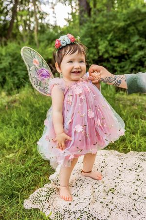 Smiling baby girl in pink fairy dress with wings and floral crown stands barefoot on lace blanket in green forest.