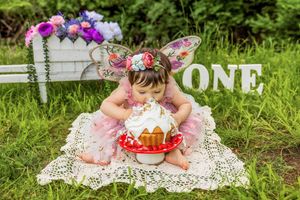 Baby girl in pink tutu and fairy wings smashes first birthday cake outdoors on lace blanket with ONE sign.
