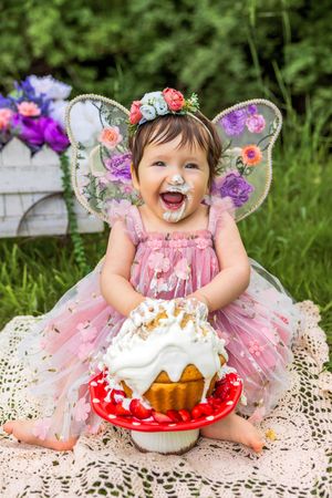 Toddler girl in pink fairy costume with butterfly wings and floral crown smashing her first birthday cake outdoors.