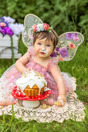 Baby girl in pink fairy costume with floral wings celebrates first birthday smashing white frosted cake outdoors.