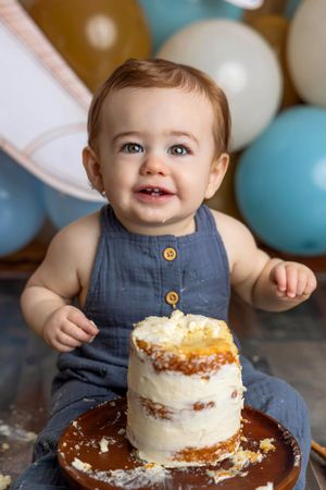 Baby in blue romper smashing first birthday cake surrounded by blue and gold balloons.