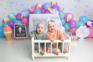 Two babies sit in a white crib surrounded by colorful balloons and ice cream decorations for a first birthday cake smash.