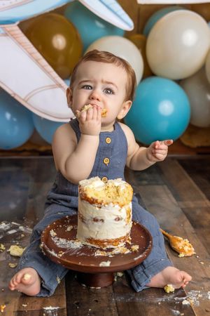 Baby in blue romper eating first birthday cake smash with balloons in background.