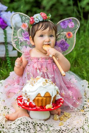 Baby girl in pink fairy costume with butterfly wings and floral crown eating cake during first birthday smash cake photoshoot.