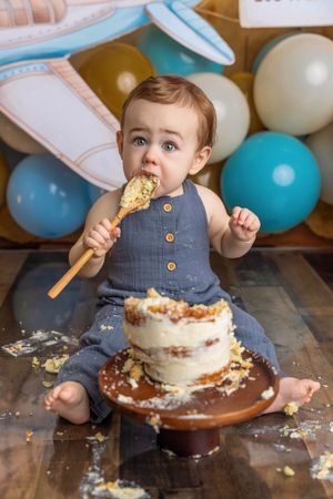 Baby boy in blue romper smashing first birthday cake with wooden spoon, surrounded by blue and gold balloons.