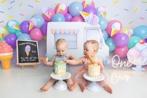 Twin babies celebrate first birthday with ice cream theme, colorful balloon arch, and individual smash cakes in a festive studio setup.