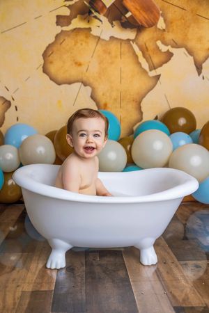Happy baby sitting in a white clawfoot bathtub surrounded by gold and blue balloons with a world map backdrop.