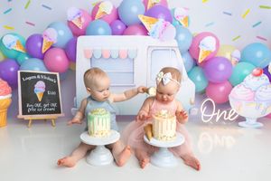 Two babies celebrate first birthday smash cake session with ice cream theme, colorful balloons, and matching white cakes.