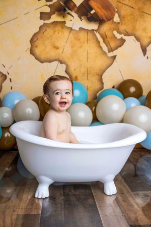 Smiling baby sitting in a white clawfoot bathtub surrounded by blue and beige balloons with a vintage world map backdrop.