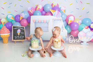 Twin babies celebrate their first birthday smashing cakes surrounded by colorful balloons and an ice cream themed backdrop.
