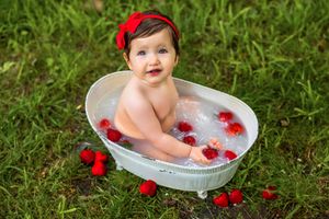 Baby girl in red headband smiling in a white mini bathtub with strawberries outdoors on green grass.