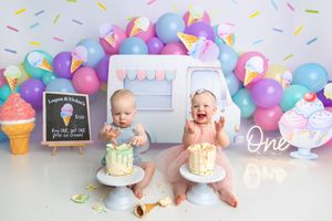 Twin babies enjoy their first birthday cake smash with ice cream theme, colorful balloons, and pastel decorations.