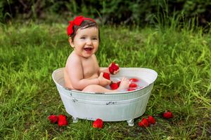 Happy baby girl sitting in a small white bathtub outdoors with strawberries and milk, wearing a red bow headband.