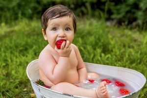 Baby sitting in a white tub outdoors eating a strawberry, surrounded by rose petals in milky water.