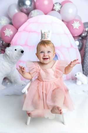 Smiling baby girl in pink tutu dress and crown sits before pink igloo backdrop with polar bears and balloons.