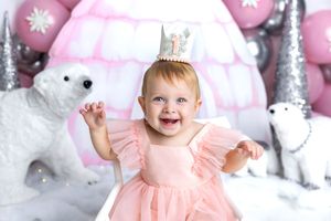Smiling baby girl in pink tutu dress and birthday crown sits among white polar bears and pink balloon decorations.