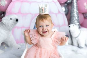 Smiling baby girl in pink dress and number 1 crown celebrates first birthday with polar bear decorations.