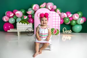 Baby girl in white dress sits on stool at watermelon-themed first birthday cake smash with pink balloons.