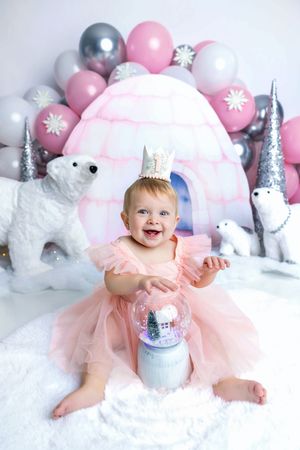 Smiling baby girl in pink tutu dress and crown hat sits with snow globe in winter wonderland birthday setup.