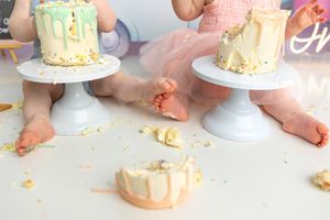 Two babies in tutus smash their first birthday cakes on white pedestals, with frosting and crumbs scattered around.