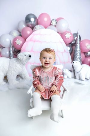 Happy baby girl in pink outfit sits on white chair surrounded by polar bears and balloons in winter birthday scene.