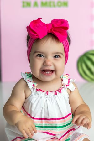 Smiling baby girl wearing a pink bow headband and striped dress, showing first teeth, sitting in front of a colorful background.