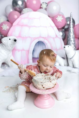 Baby girl in pink dress smashing first birthday cake at winter wonderland themed party with igloo backdrop.
