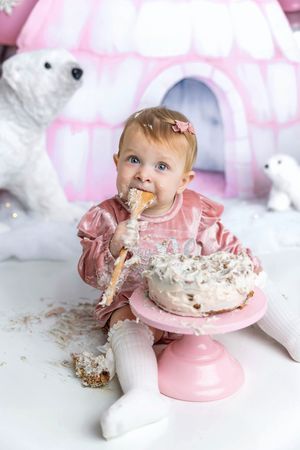 Baby girl in pink dress eating smash cake at first birthday party with polar bear igloo decorations.