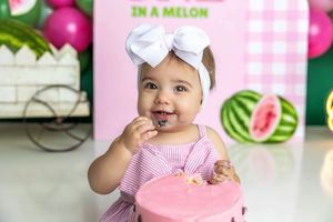 Baby girl in pink dress and white bow headband smiles while eating pink cake at watermelon-themed first birthday party.