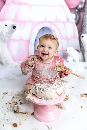 Happy baby girl in pink dress smashes first birthday cake on pink cake stand with polar bear igloo backdrop.