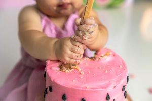 Baby girl in pink dress smashing a pink watermelon-themed first birthday cake with a candle.