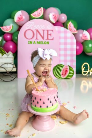 Baby girl celebrates first birthday smashing a watermelon-themed cake at a ONE in a Melon party backdrop.