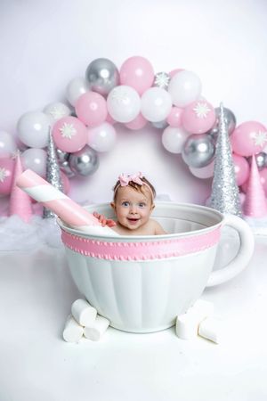 Baby girl sitting in giant teacup with pink balloons and marshmallows in a whimsical winter wonderland photo setup.