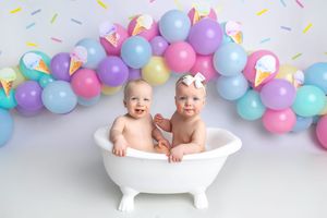 Two babies sit in a white mini bathtub surrounded by colorful balloons and ice cream decorations.