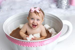 Smiling baby girl with pink bow headband sitting in oversized teacup with hot chocolate and marshmallows.