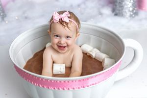 Smiling baby girl wearing pink bow headband sitting in oversized decorative teacup filled with chocolate milk and marshmallows.