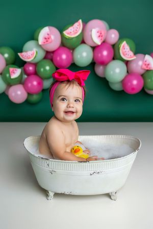 Smiling baby in white tub with rubber duck, pink bow headband, watermelon balloon backdrop.