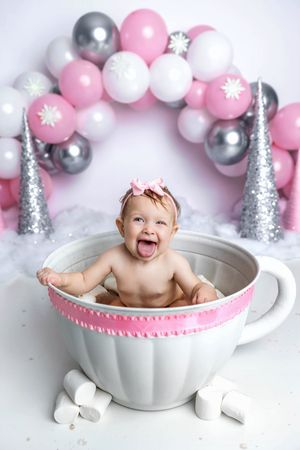 Laughing baby sits in giant teacup prop surrounded by marshmallows with pink and silver balloon arch backdrop.